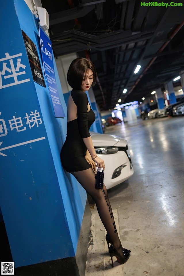 A woman in a black dress leaning against a blue wall.