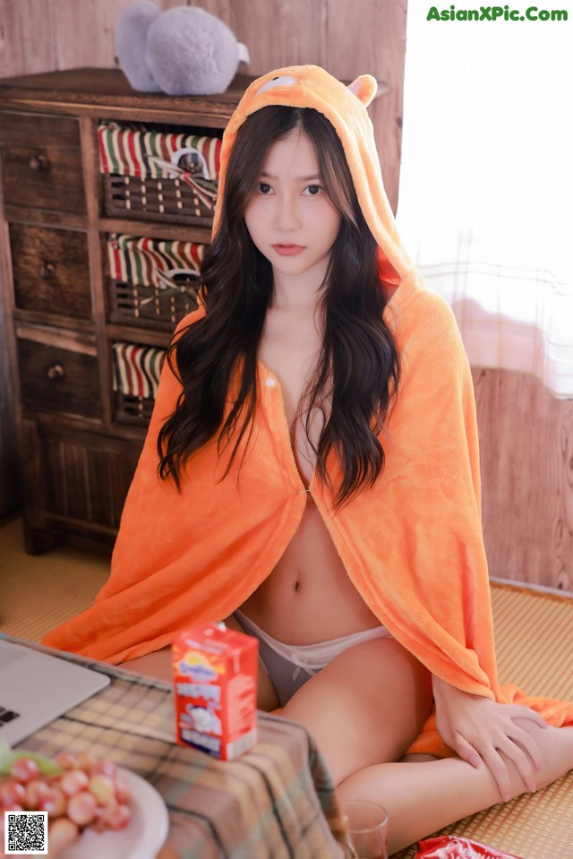 A woman in an orange hooded towel sitting on a bed.